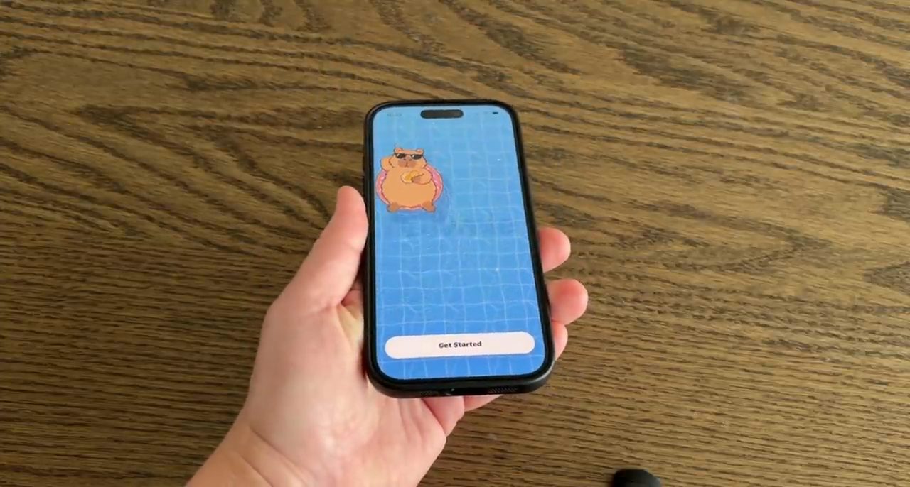 Tay cầm iPhone đang mở màn hình hồ bơi với capybara trên phao donut và nút Get Started